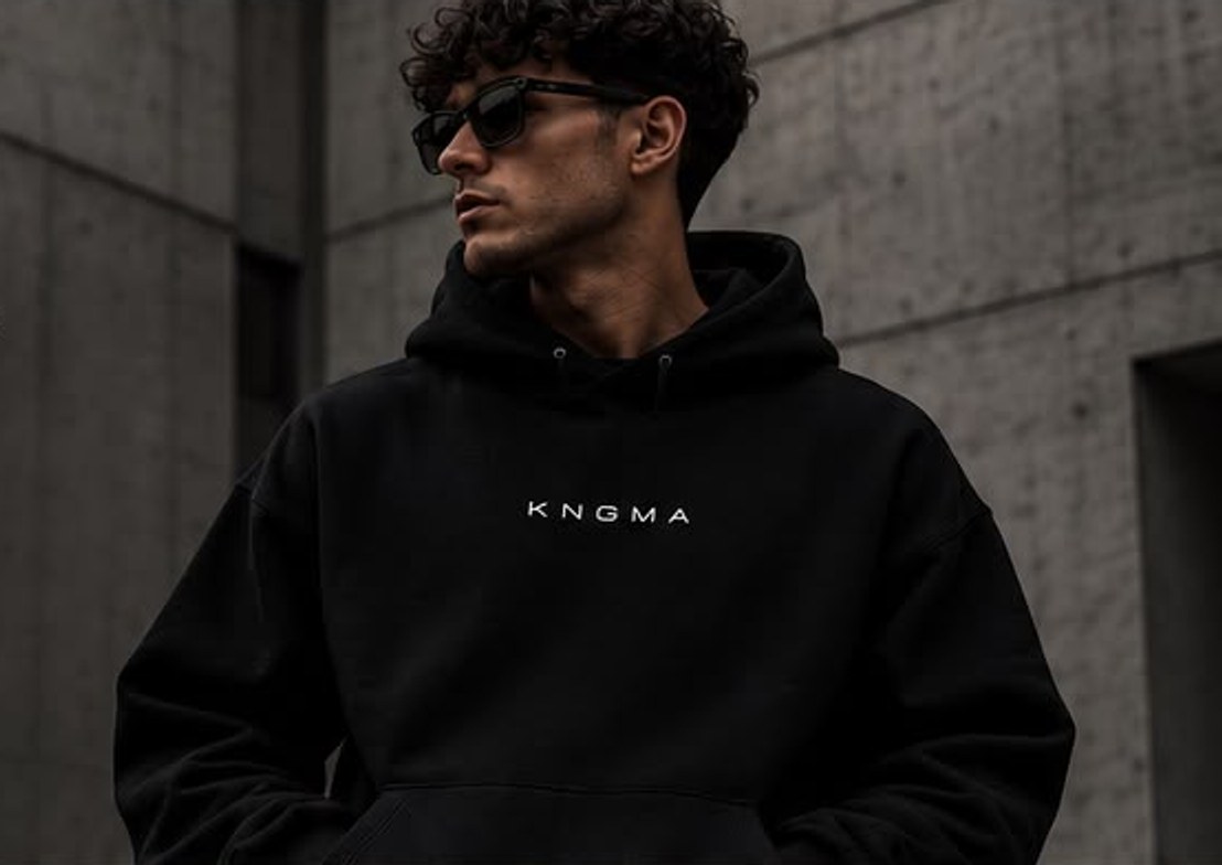 Man met zonnebril in een zwarte KNGMA-hoodie voor een betonnen muur