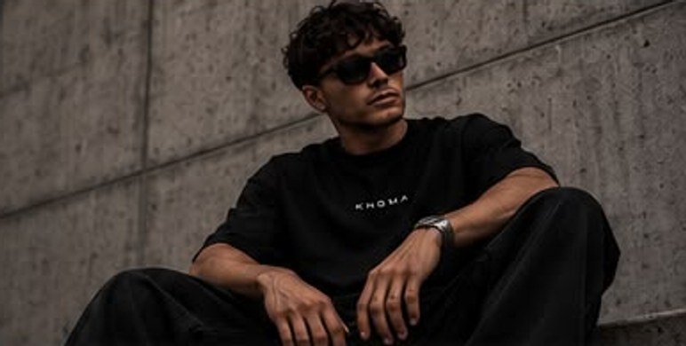 Zittende man in een zwart KNGMA-shirt tegen een betonnen muur